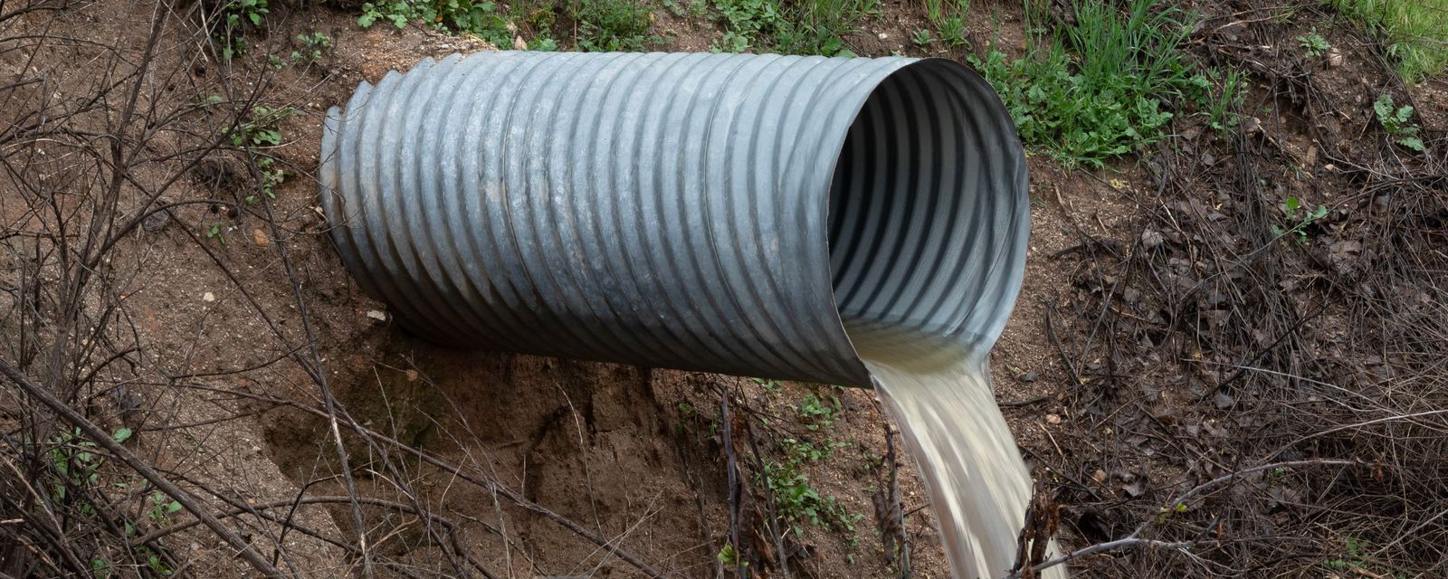 Muddy water pours from the mouth of a corrugated metal culvert pipe set in an eroded dirt bank.