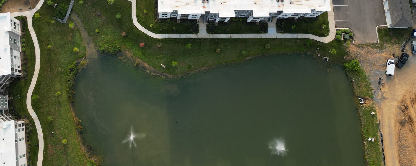 Aerial view looking straight down at a green pond with two fountains, bordered by an apartment building, a curving walkway and a strip of bare graded soil along the right edge.
