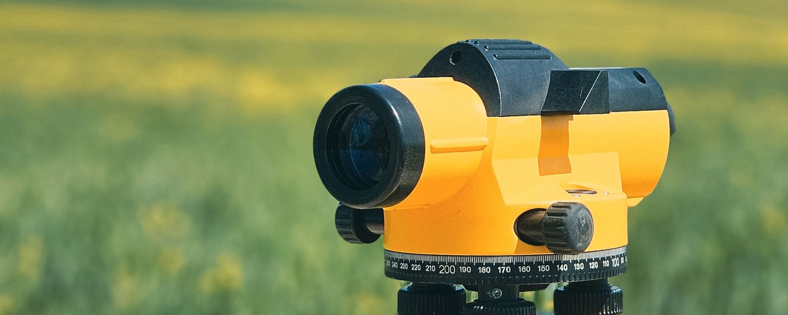 A yellow automatic surveying level set up in a grassy field with blurred yellow wildflowers behind it.