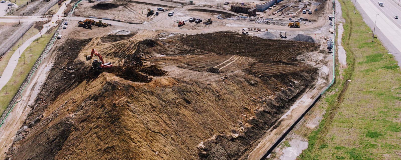 Aerial view of a large graded construction site: an excavator works on a soil stockpile, and a fence lined with dark fabric separates the bare earth from a strip of grass beside a highway.