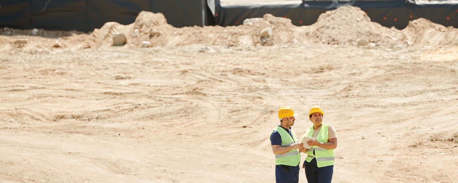 Two workers in yellow hard hats and high-visibility vests look at a tablet on a graded dirt construction site, with soil stockpiles and black perimeter sheeting behind them.