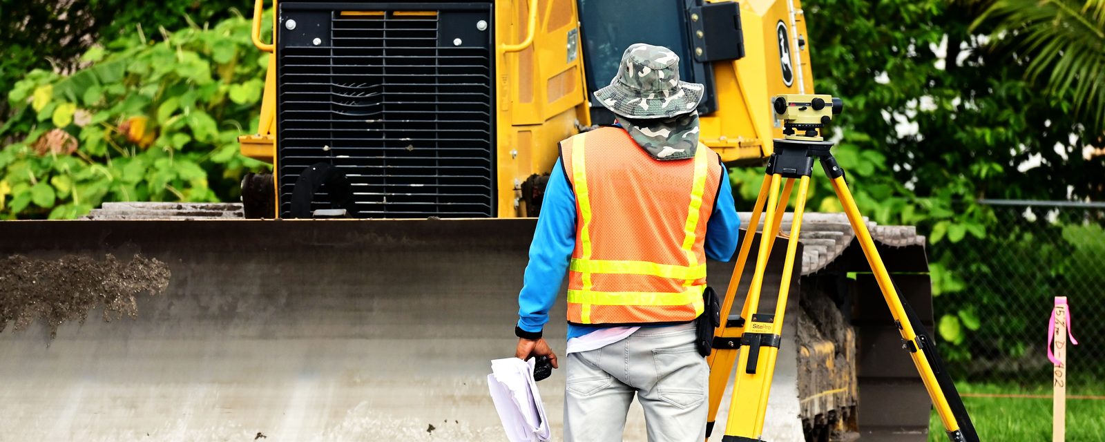 A surveyor in a hi-vis vest stands beside an automatic level on a yellow tripod on a graded dirt site, with a bulldozer blade behind him.