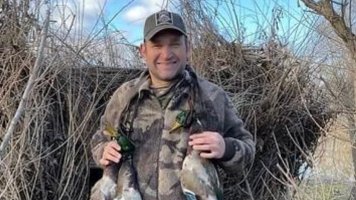 Chris Duffy in a camouflage jacket and cap, holding two ducks in front of a brush blind